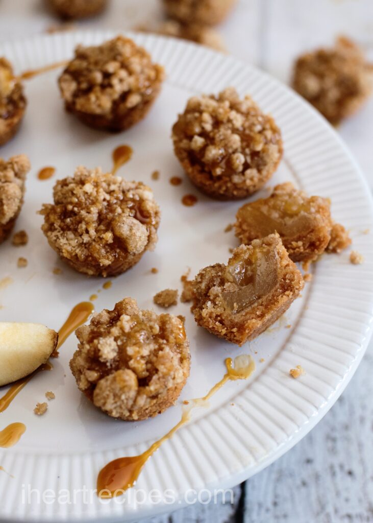 A few mini caramel apple crisps are arranged on a white platter and drizzled with caramel. One crisp is bitten in half revealing a tender apple filling. 