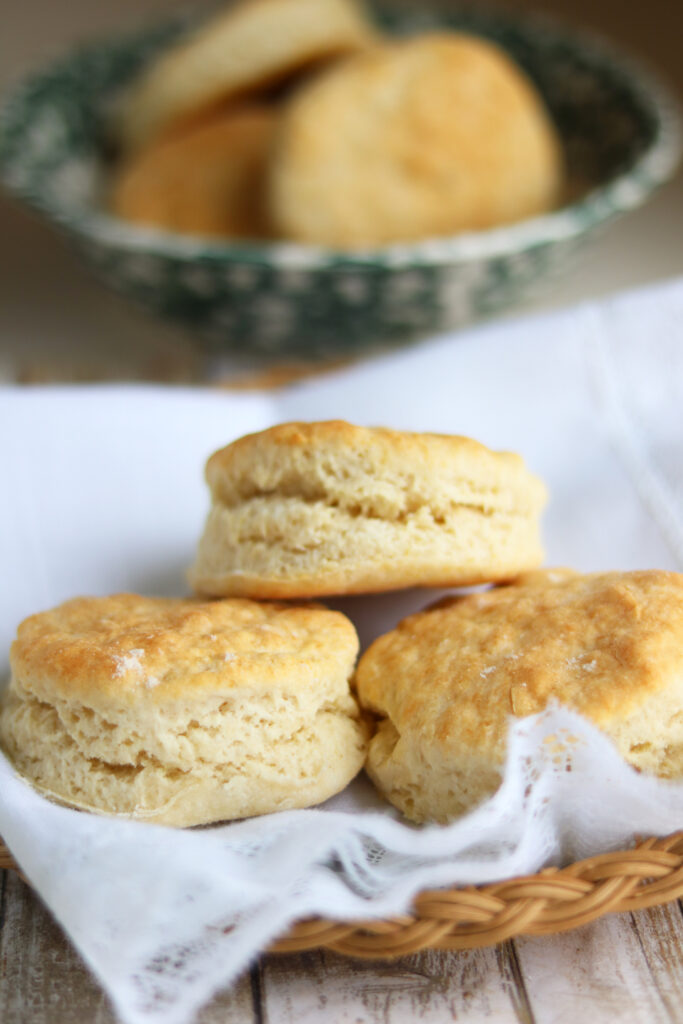 Three golden buttermilk biscuits stacked on a white cloth-lined basket. A bowl of biscuits is seen in the background. 