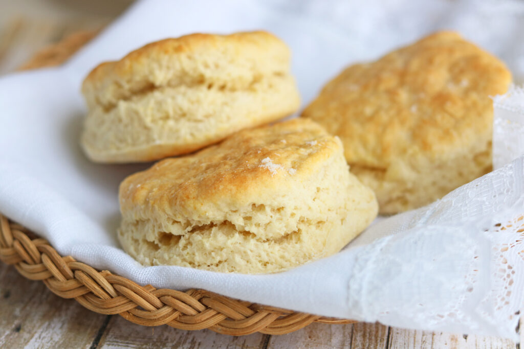 Three golden and moist homemade buttermilk biscuits resting in a wicker basket lined with a white cloth.  