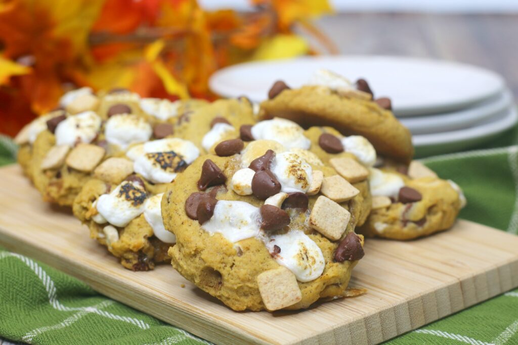 A few golden Pumpkin S'mores Cookies are stacked on on a wooden board.  