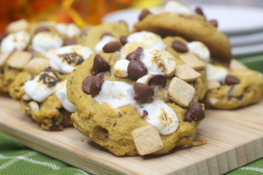 Pumpkin S'mores Cookies stacked on a wooden board. Soft chocolate chips and melted marshmallows dot the golden cookies. 