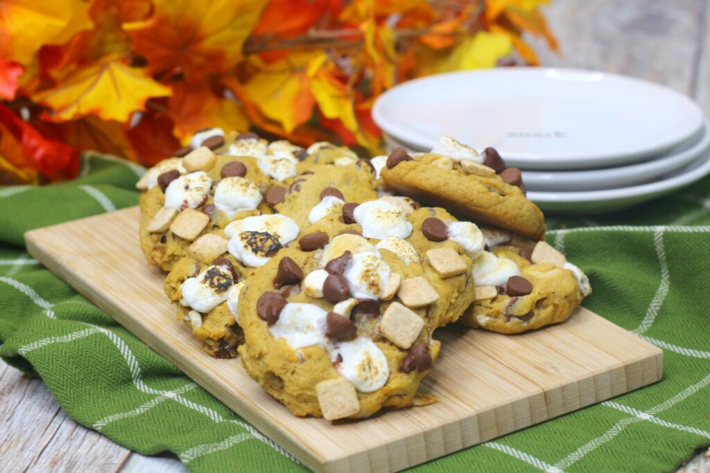 Golden Pumpkin S'mores Cookies arranged on a wood serving platter. Melted marshmallows, soft chocolate chips and s'mores baking chips dot the tops. 