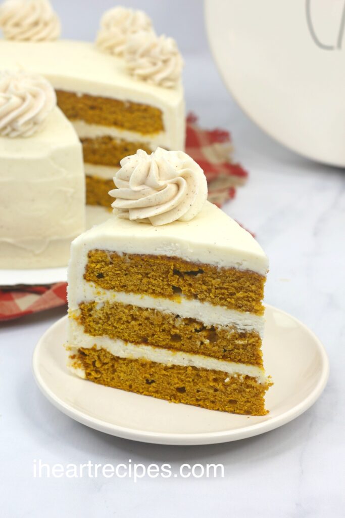 A slice of three-layered pumpkin cake topped with smooth cream cheese frosting. The rest of the moist layered pumpkin cake is in the background. 