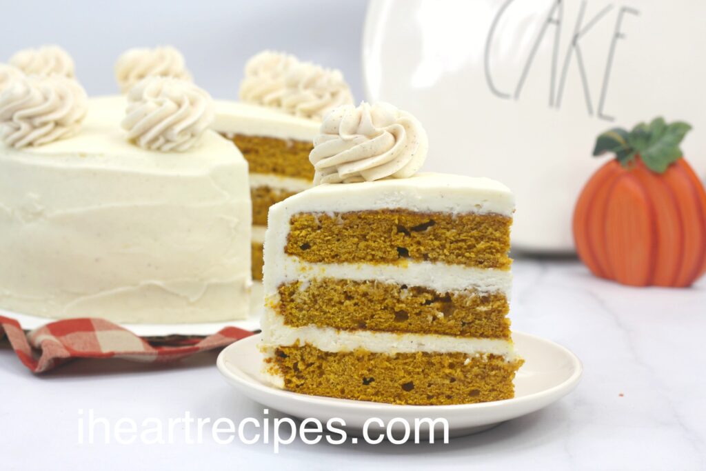 A large slice of moist layered pumpkin cake on a small white plate. It's topped with a swirl of cinnamon cream cheese frosting. 