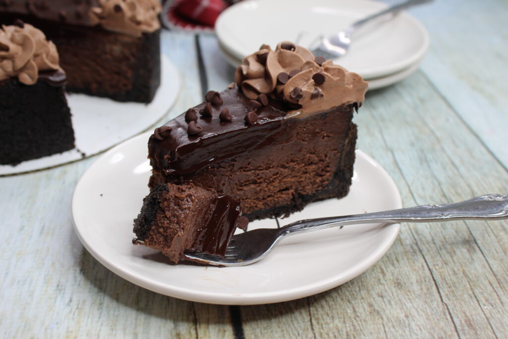 A slice of chocolate cheesecake on a round white plate. A forkful of cheesecake rests on the plate. 