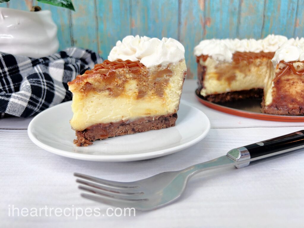 A slice of Dulce de Leche Cheesecake on a round white plate. The remaining cheesecake sits in the background. A silver and black fork is in the foreground. 