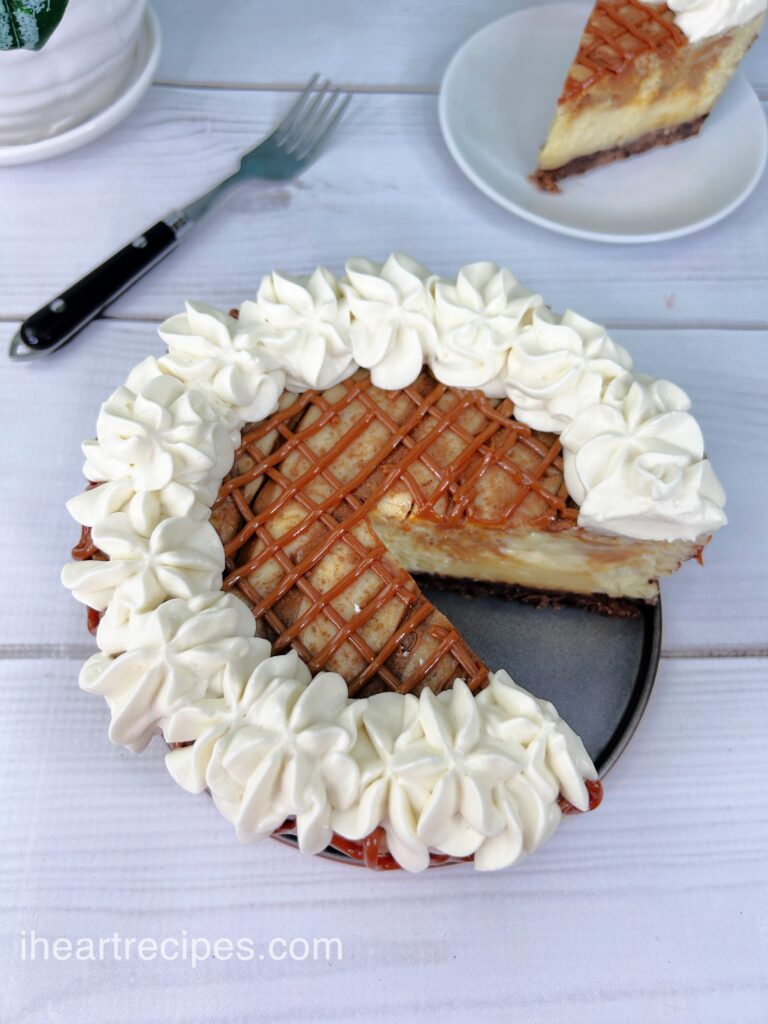 A dulce de leche cheesecake set on a white wooden table. A slice is missing from the pan and is seen in the background. 