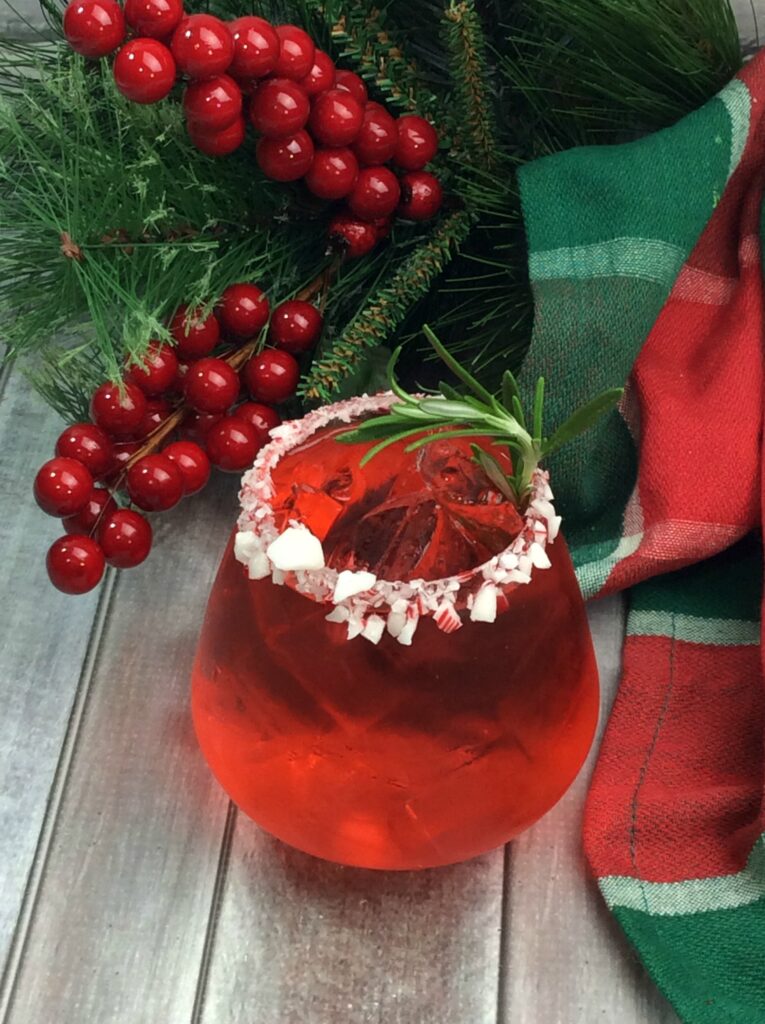 A festive red candy cane cocktail served in a glass tumbler rimmed with sweet crushed candy canes and finished off with a fresh sprig of rosemary. 