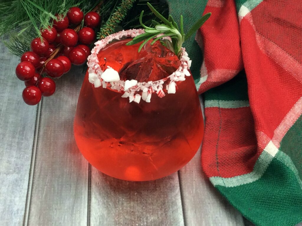 A bright red Jolly Candy Cane Cocktail served in a glass tumbler rimmed with crushed candy canes and garnished with a sprig of rosemary. 