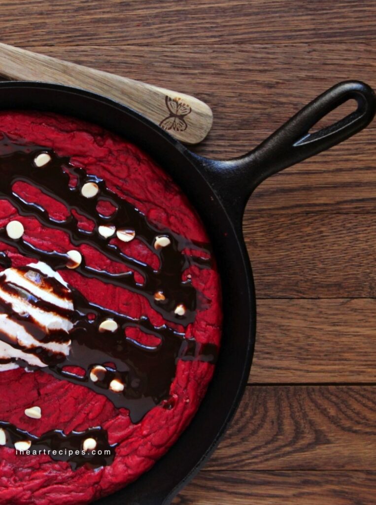 A skillet red velvet cookie served in a cast iron skillet and topped with ice cream, white chocolate chips and a chocolate sauce. 