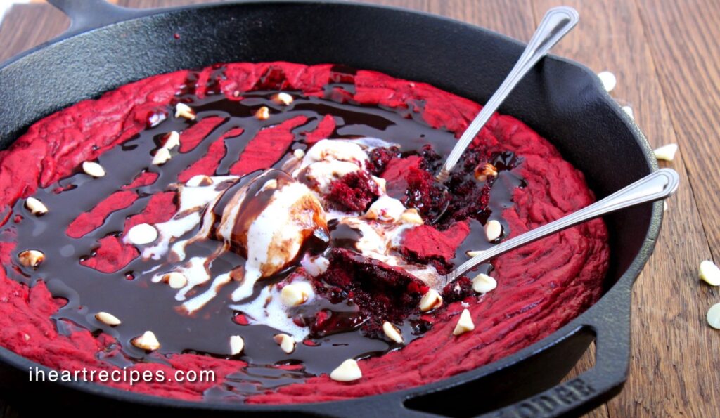 Two silver spoons dug into a sweet skillet red velvet cookie served in a cast iron skillet. 