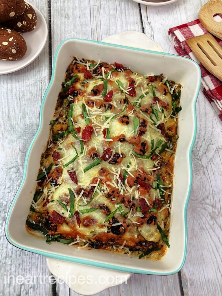 A colorful Baked Tuscan Chicken Casserole in a turquoise and white dish. Fresh rolls and wooden serving utensils lay nearby.  