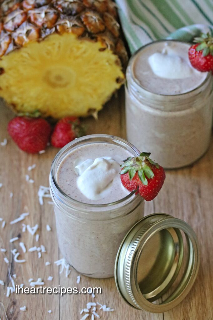 Pineapple Strawberry Coconut Smoothie fills a clear mason jar. It's garnished with a fresh strawberry and a dollop of coconut whip. The lid and ring rest against the side of the jar. 