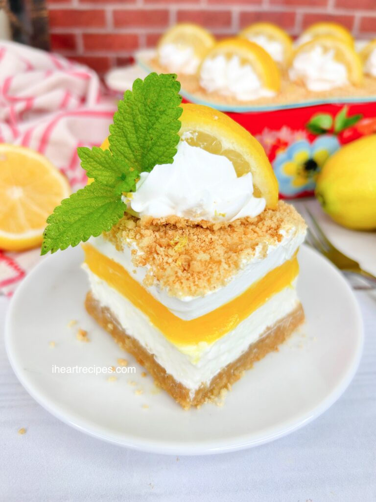 A serving of Layered Lemon Dream Dessert served on a white plate. The remaining cake is in a colorful bake dish which is seen in the background. 