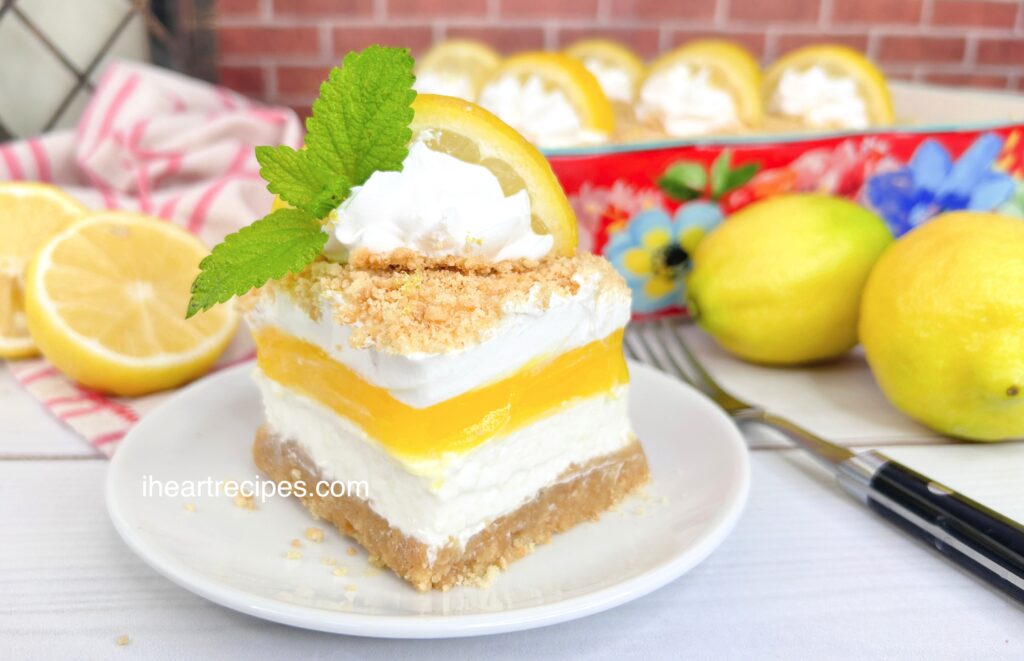 A square of Layered Lemon Dream Dessert garnished with a dollop of whipped cream, a lemon slice and a sprig of mint, all served on a round white plate. 