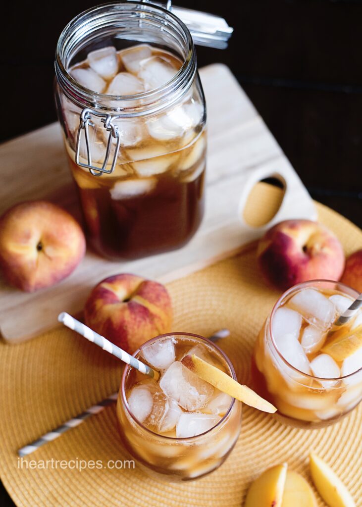 Two glasses filled with Southern Peach Iced Tea and garnished with a slice of fresh peach. A glass jar filled with tea and ice sits nearby alongside fresh peaches. 