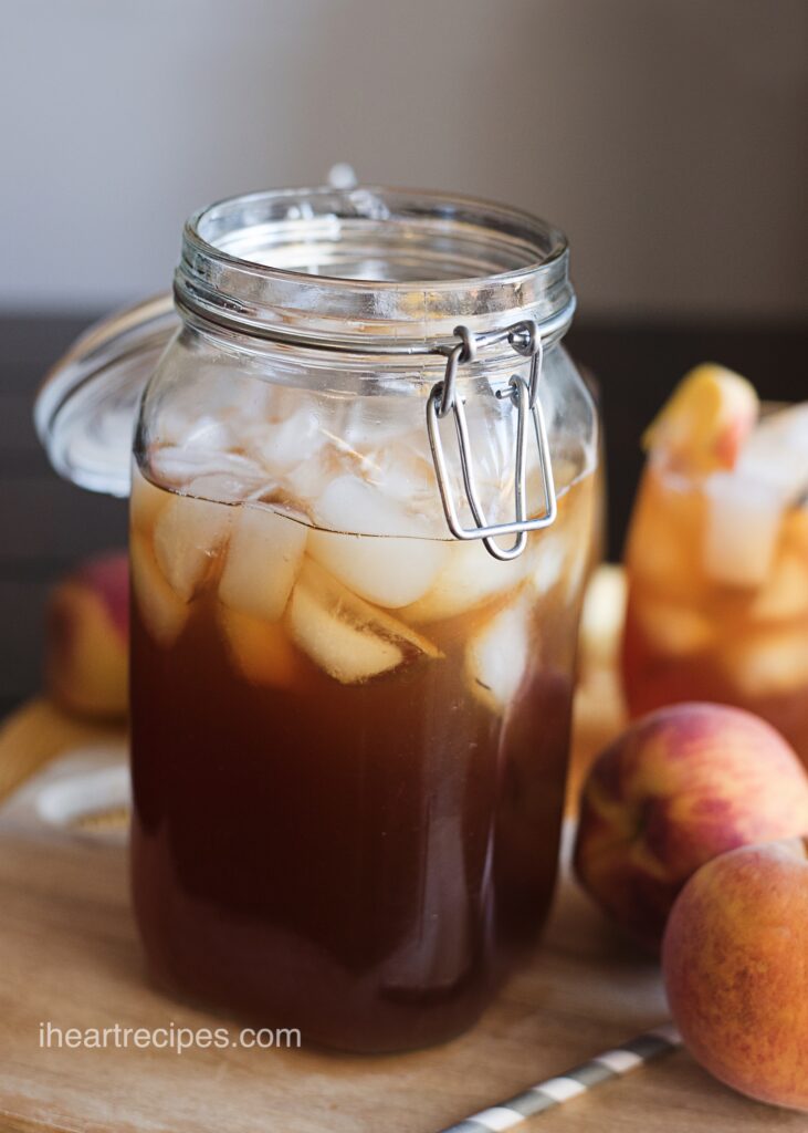 A large glass jar filled with Southern Peach Iced Tea and ice. Fresh peaches lay nearby. 