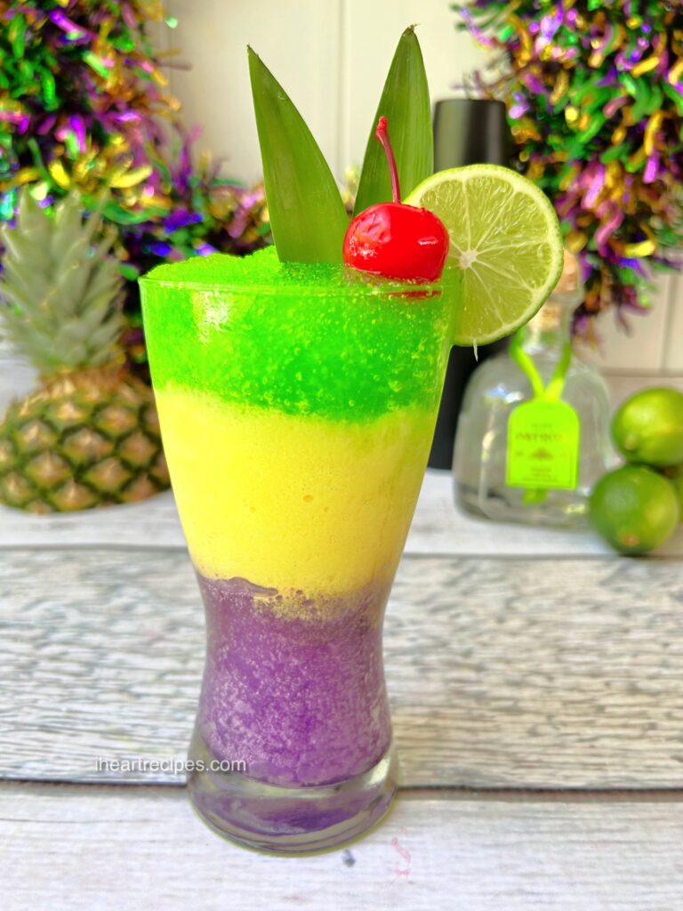 A colorful frozen Mardi Gras Margarita served in a tall glass garnished with pineapple leaves, a lime slice and a cherry. 