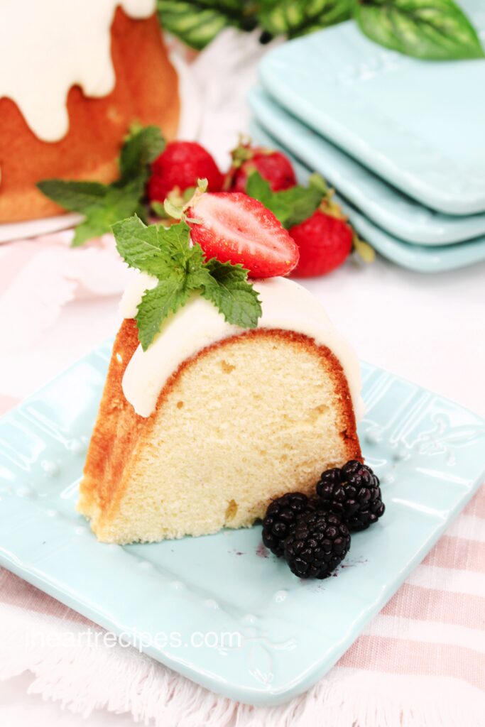 A sliced strawberry and fresh mint leaves sit atop a frosted slice of pound cake on a pale blue square plate.