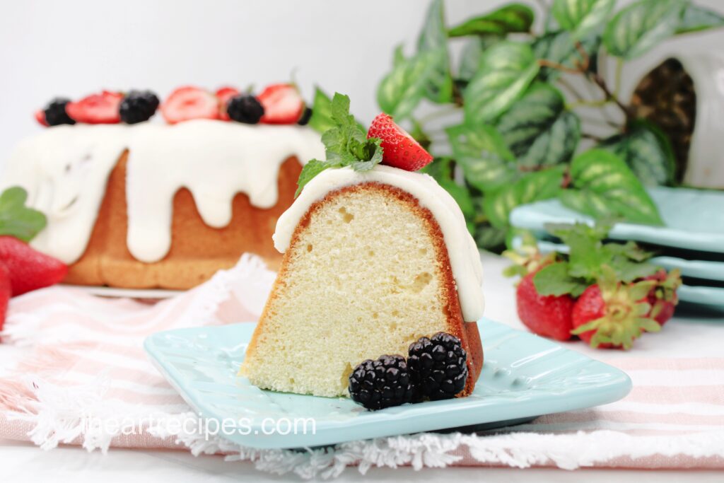 A slice of Sweet White Wine Pound Cake frosted with thick white icing and garnished with fresh berries and mint leaves. The remaining cake sits in the background. 
