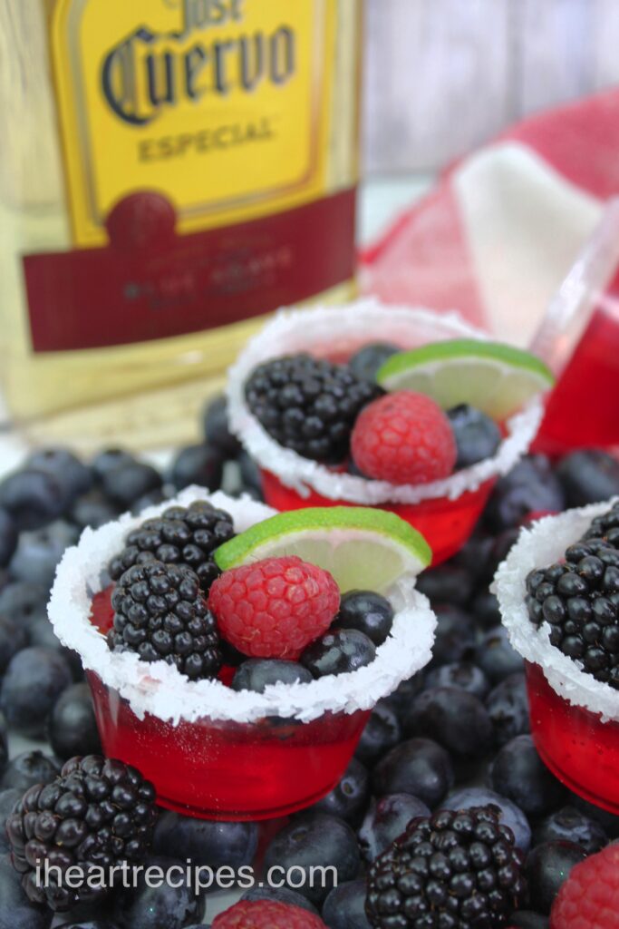 Bright red Tri Berry Jello Shots arranged on a bed of fresh berries. A bottle of Jose Cuervo tequila is seen in the background. 