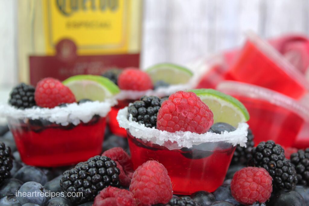 Several Tri Berry Jello Shots rimmed with salt arranged on a bed of fresh berries. A bottle of tequila is in the background. 