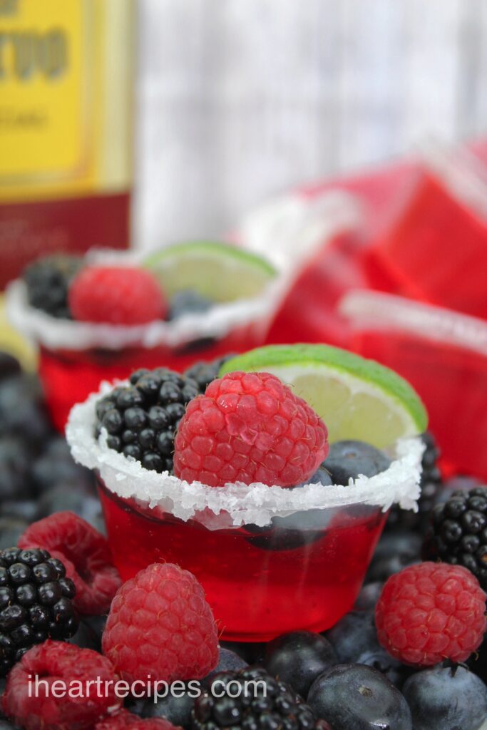 A close up image of a bright red jello shot garnished with a variety of fresh berries and a slice of lime. 