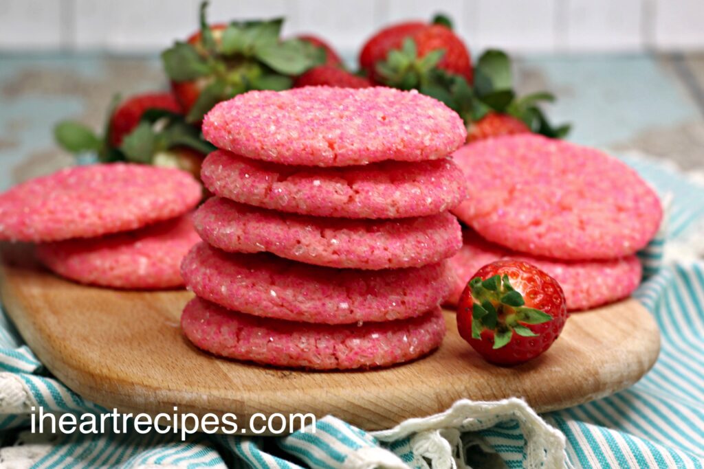 A stack of Strawberry and Champagne Sugar Cookies on a wooden serving platte. Fresh strawberries lay nearby. 