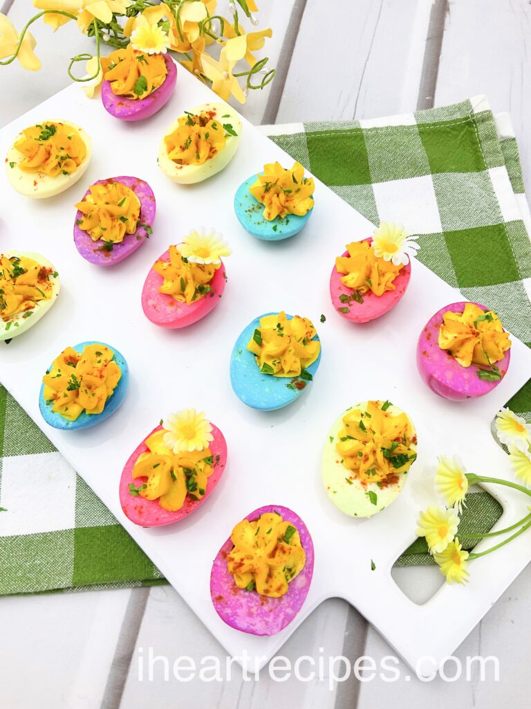 Pastel dyed deviled eggs on a white serving board set on a green and white gingham cloth. 