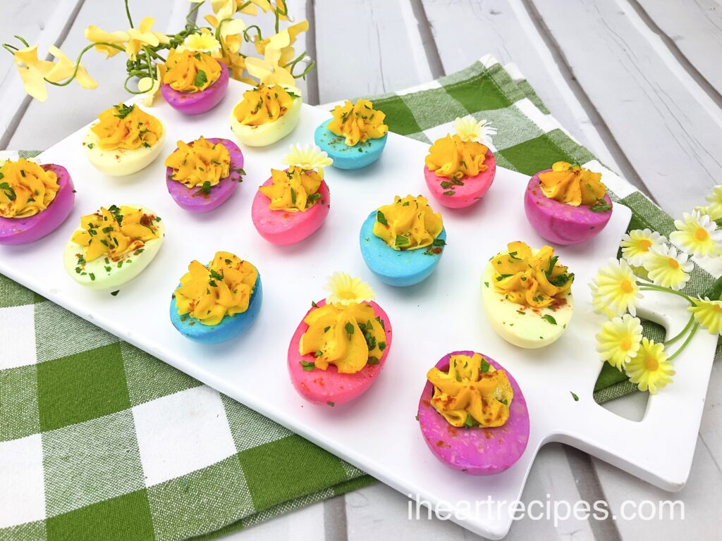 A white serving board with rows of pastel colored Easter deviled eggs piped with golden filling and garnished with fresh herbs and flowers. Sprigs of yellow flowers sit nearby. 