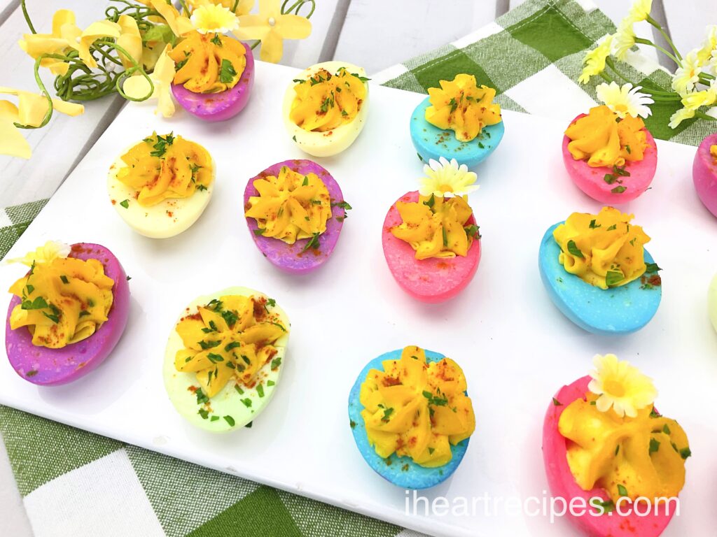 Rows of pastel colored Easter Classic Deviled Eggs filled with golden filling and garnished with fresh parsley and flowers. 