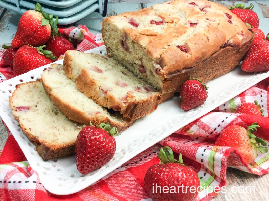 A golden loaf of strawberries and cream bread sliced on an elegant white serving platter. Whole fresh strawberries lay about.  
