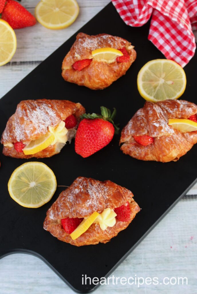 Four strawberry & lemon custard stuffed croissants set on a black board. Fresh strawberries and lemon slices lay nearby.