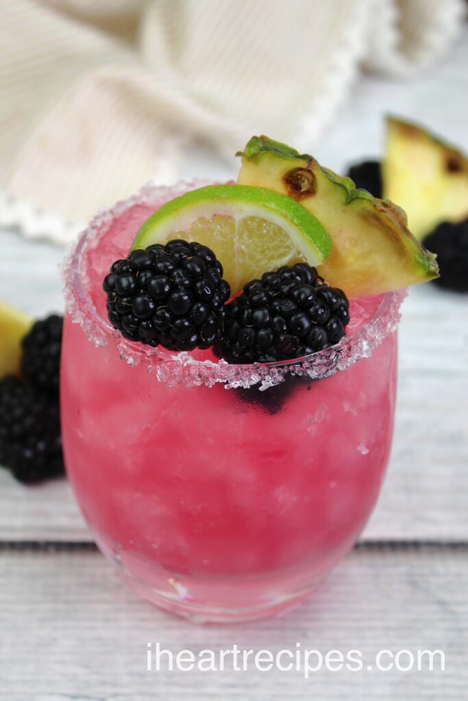 Fresh blackberries and slices of lime and pineapple garnish a bright pink drink in a clear glass tumbler rimmed with salt.