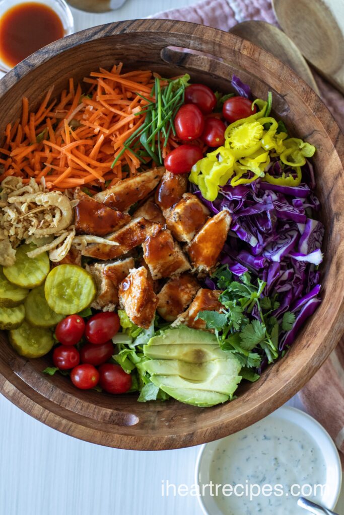 A wooden bowl filled with BBQ Chicken Salad. Bright veggies, creamy avocado, crisp onion shreds and savory chicken are seen. 