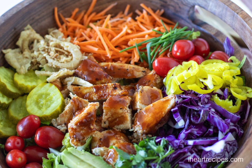 A wooden bowl filled with a rainbow of vegetables and pieces of tender BBQ Chicken. 