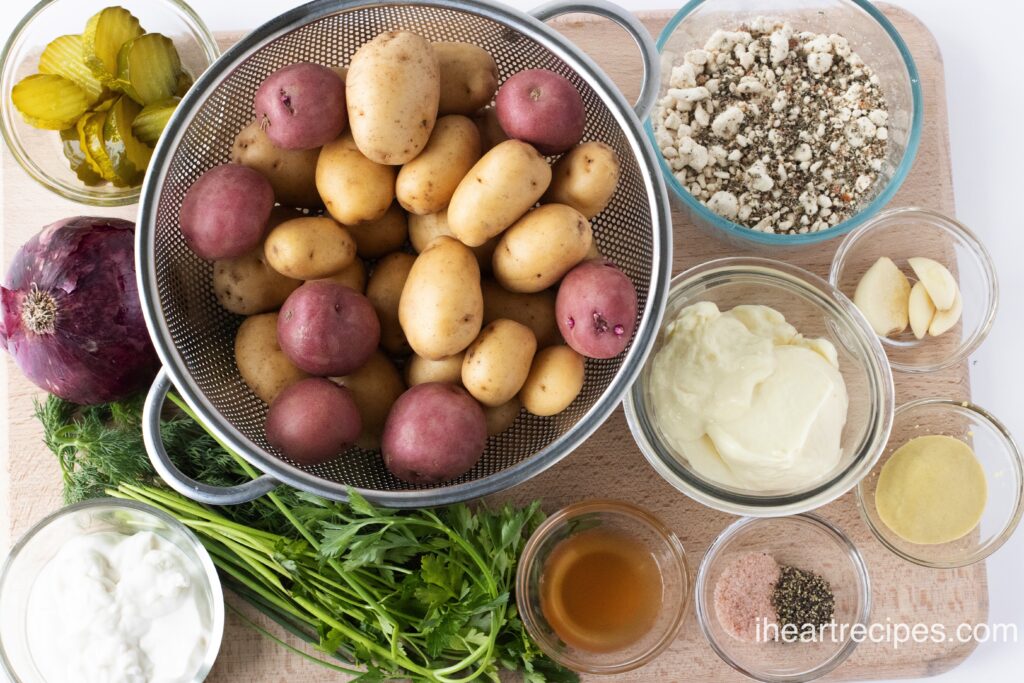 All of the ingredients for smashed potato salad are separated into individual glass bowls set on a wood cutting board.