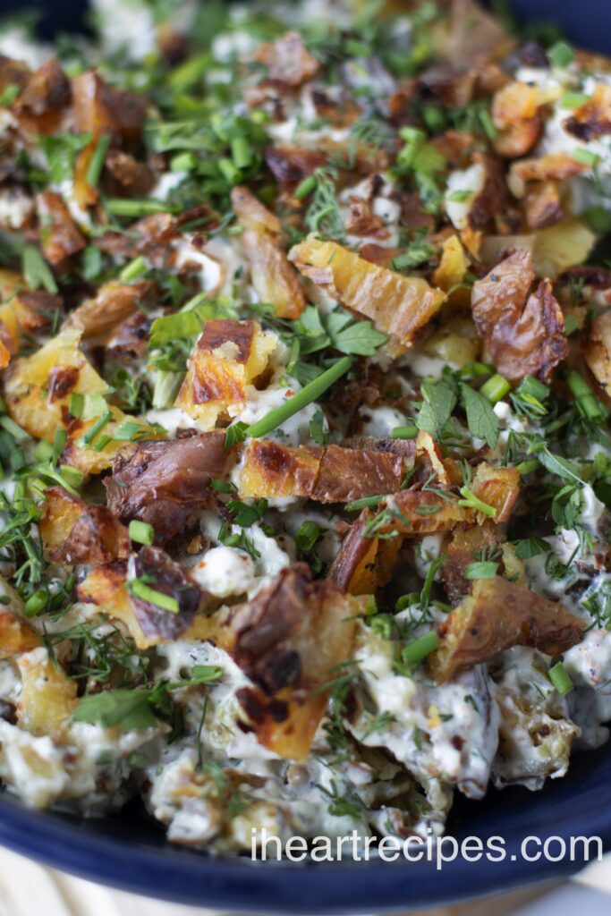 A close up image of smashed potato salad. Bits of golden potato, creamy dressing and cheese along with fresh herbs are seen. 