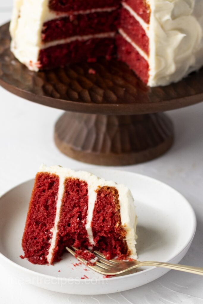 Three Layer Southern Red Velvet Cake on a stand, with a slice of cake on a small plate.